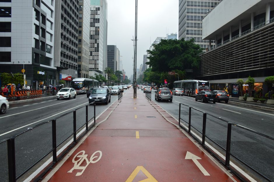 Foto ilustrativa de avenida com ciclovia vermelha no canteiro central e prédios comerciais dos dois lados
