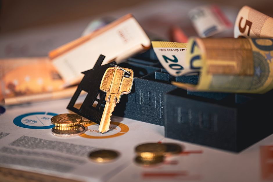 Foto ilustrativa de chave com chaveiro de casa, moedas e cédulas sobre papéis com gráficos